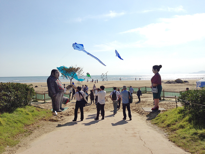 Main Dadaepo Beach entrance overlooking the 2015 Sea Art Festival (SAF). Greeting visitors are two figurative sculptures by WON-GEUN KIM, characters that reminded the artist of two mysterious guests that arrived at the artist’s childhood home when he was little, whose presence caused his parents to fight after their departure. He never found out who they were. Flying overhead are kites of sea creatures by PETER LYNN KITES, a SAF special exhibition.